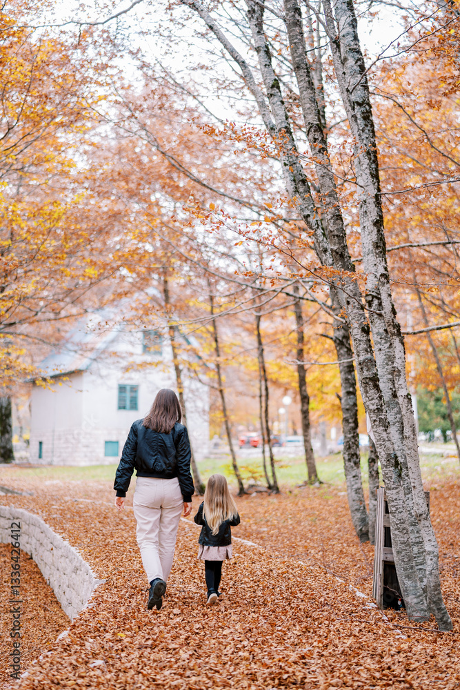Fototapeta premium Mother and a little girl walk along a path in an autumn park towards the house. Back view