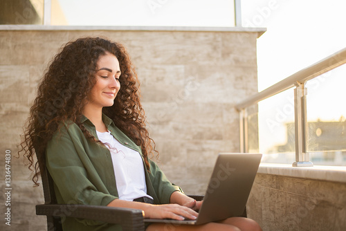 Beautiful curly woman working on laptop sitting on balcony at sunset, warm sun rays. Work on the Internet, online shopping, communication on social networks, online learning, freelancer, remote work