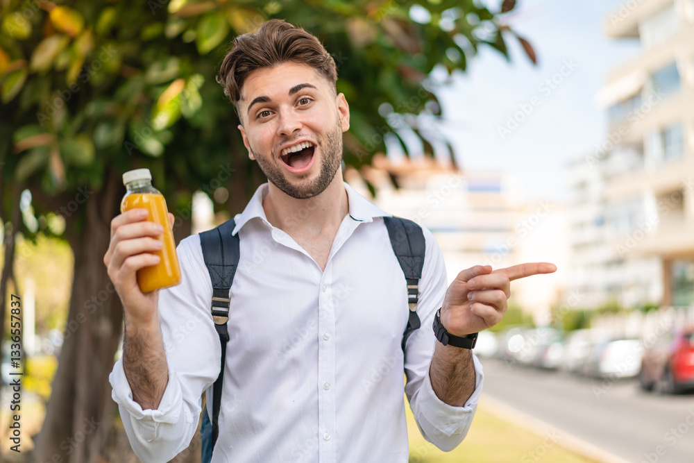 Young handsome man holding an orange juice at outdoors surprised and pointing finger to the side Young handsome man holding an orange juice at outdoors surprised and pointing finger to the side