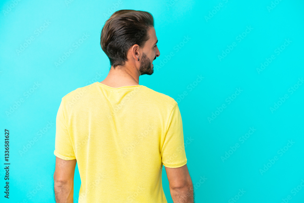 Young caucasian man isolated on blue background in back position and looking side Young caucasian man isolated on blue background in back position and looking side