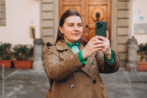 Picture of pretty young woman holding phone in hands on the street. Redhead girl making selfie photo on urban background. Lifestyle, travel, tourism, technology, blogging, communication concept.