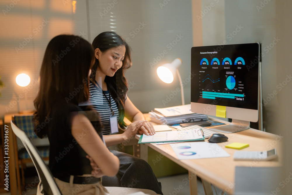 Asian businesswomen reviewing marketing data displayed on computer screen in modern office at night Asian businesswomen reviewing marketing data displayed on computer screen in modern office at night