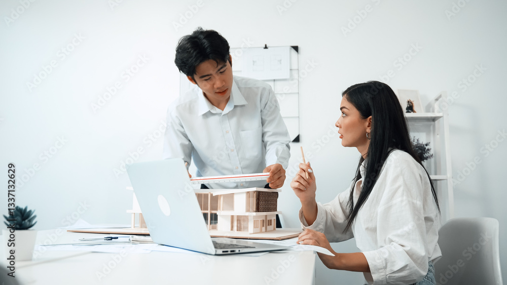 Professional male asian architect using ruler to measure house model length while young beautiful caucasian colleague using laptop to analyzed data on meeting table with house model. Immaculate. Professional male asian architect using ruler to measure house model length while young beautiful caucasian colleague using laptop to analyzed data on meeting table with house model. Immaculate.