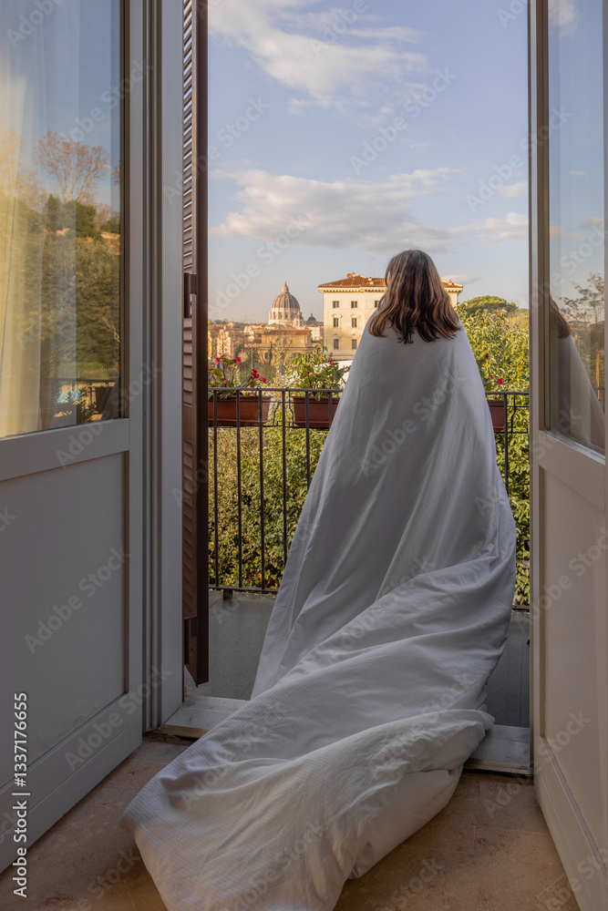 A woman wrapped in a white blanket stands on a cozy balcony, gazing at the stunning morning view of St. Peters Basilica in Vatican City