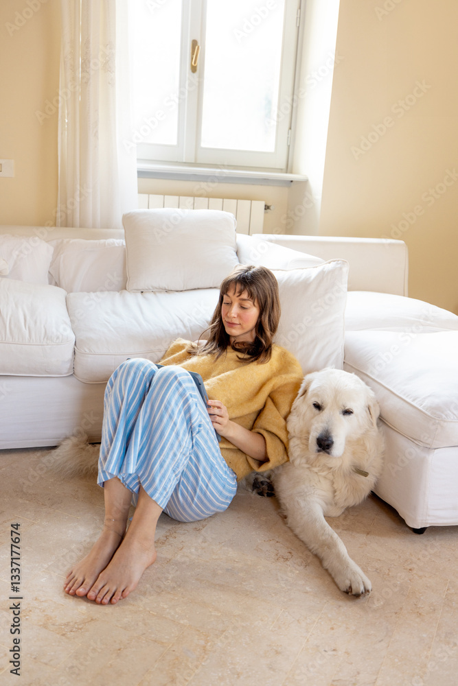 Relaxed woman in a warm sweater and striped pajama pants lounges on a soft carpet, cuddling her fluffy white dog while reading a book. A peaceful moment of comfort and companionship at home