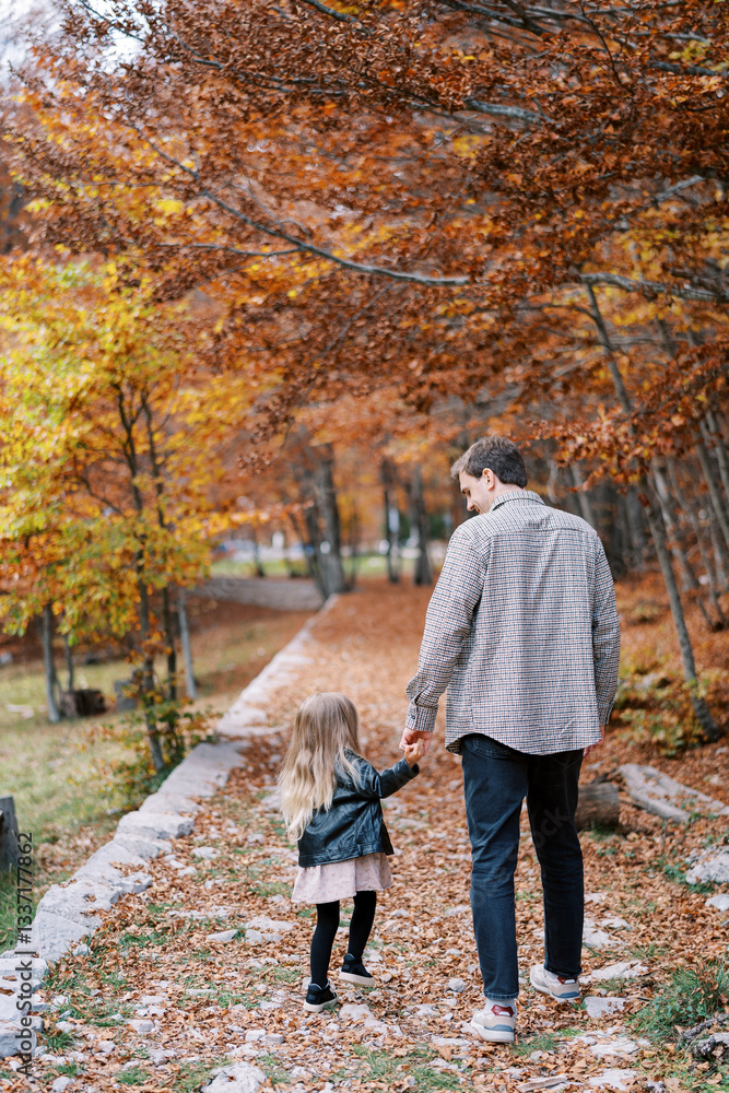 Fototapeta premium Dad and a little girl are walking holding hands along a rocky path in the autumn forest. Back view