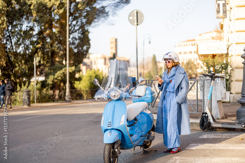 Obraz na płótnie A fashionable woman in a blue coat and jumpsuit stands by her scooter, checking her phone on a sunny day in Rome, Italy. A modern take on urban elegance, independence, and city lifestyle w salonie