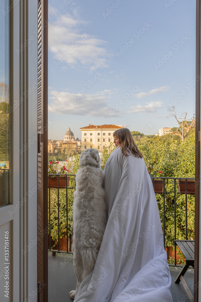 A woman wrapped in a white blanket stands on a balcony with her large fluffy dog, gazing at the Vatican dome in the distance
