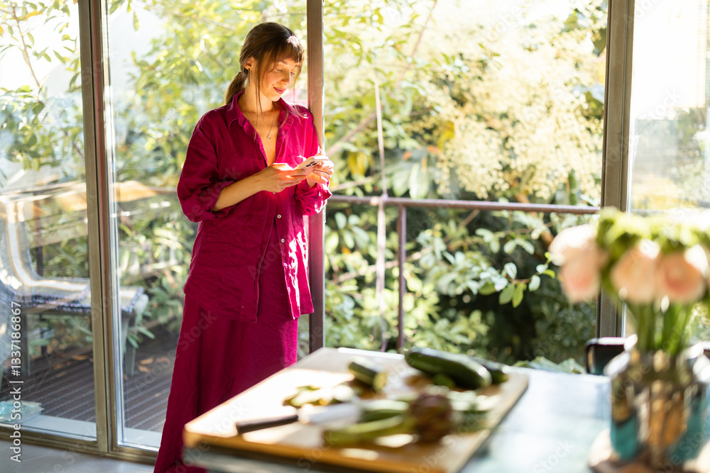 A woman in cozy pajamas enjoys a quiet morning in a sunlit kitchen, checking her phone. The warm natural light and greenery create a peaceful, slow-living atmosphere