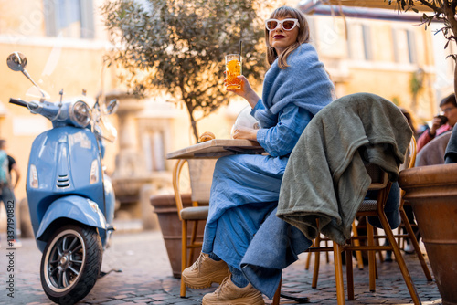 Obraz na płótnie A fashionable Italian woman sits at a cozy outdoor cafe in Rome, sipping an aperitivo. Her scooter is parked nearby, adding to the effortless elegance of Italian city life w salonie