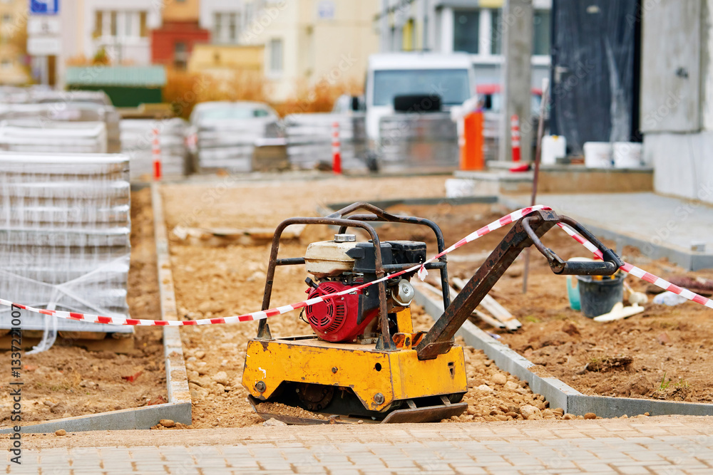 Plate compactor stationed on sandy ground. Road construction site with yellow plate compactor, enclosed with red and white caution tape, surrounded by bricks, paving blocks for sidewalk installation