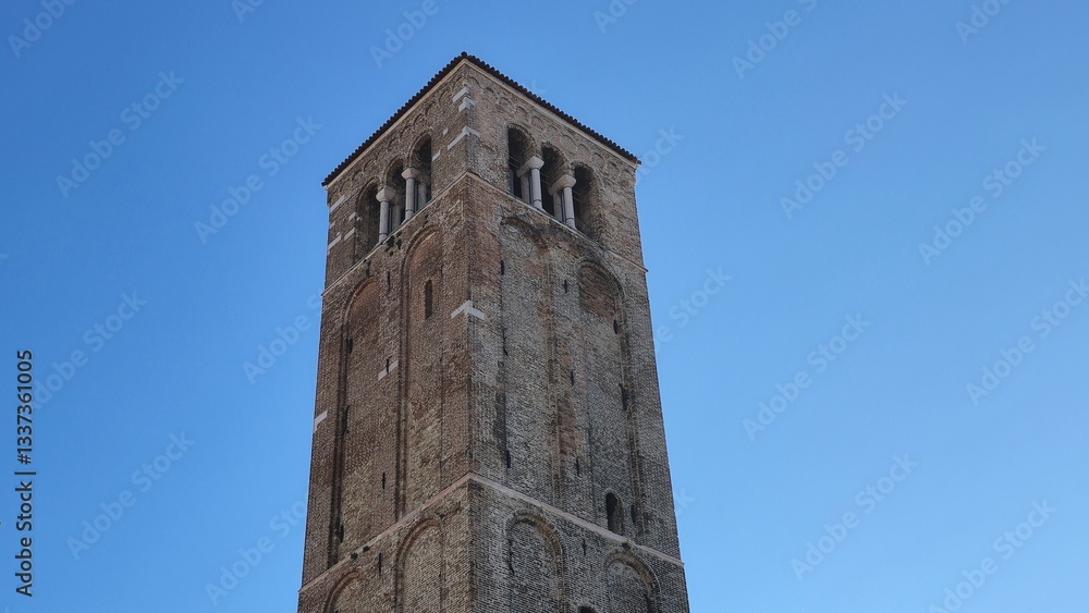 A historic brick bell tower with arched openings and decorative details stands tall against a clear blue sky. Its weathered facade and sturdy structure reflect its age and architectural significance. A historic brick bell tower with arched openings and decorative details stands tall against a clear blue sky. Its weathered facade and sturdy structure reflect its age and architectural significance.