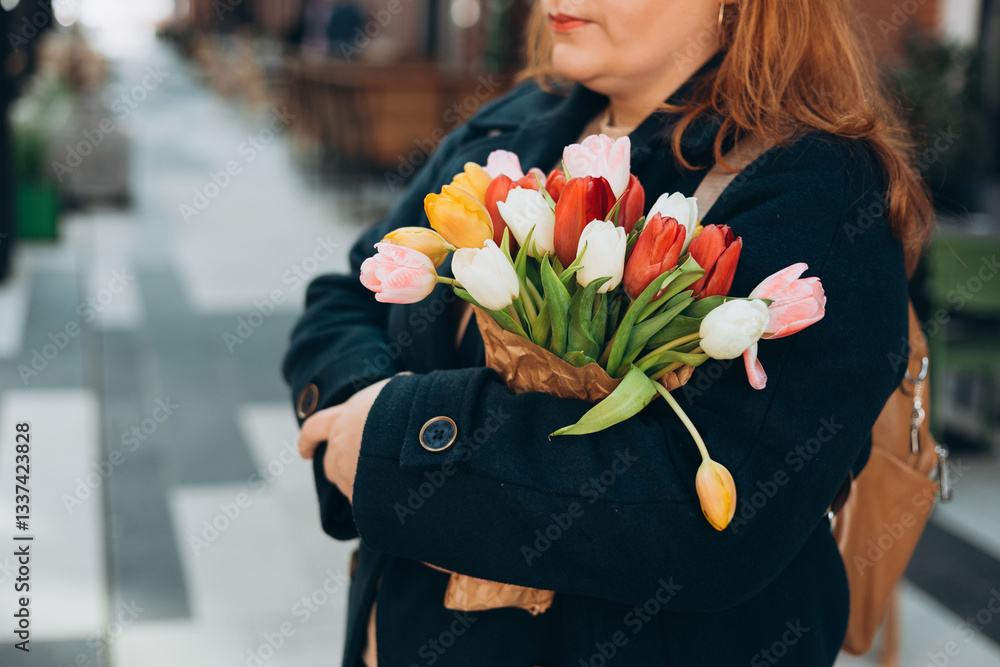 Beautiful young woman with spring tulips flowers bouquet walking at city street. Happy girl posing outdoors. Spring portrait of pretty female on urban background. High quality photo.