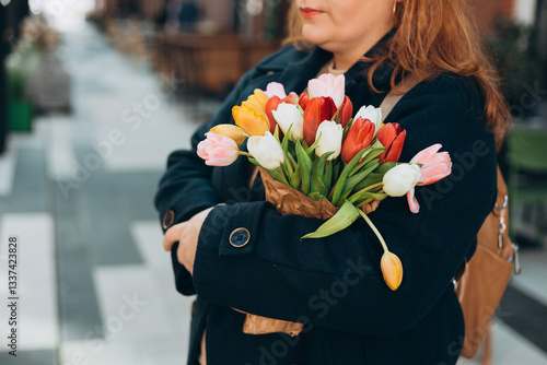 Obraz na płótnie Beautiful young woman with spring tulips flowers bouquet walking at city street. Happy girl posing outdoors. Spring portrait of pretty female on urban background. High quality photo. w salonie