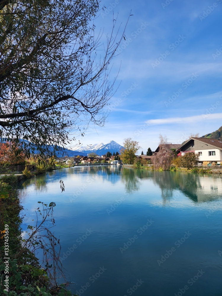 Fototapeta premium A serene river flows through a picturesque town, framed by greenery and snow-capped mountains under a vibrant blue sky