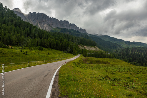 Obraz na płótnie panoramica su un ambiente di alta montagna nell'Italia settentrionale, in Veneto, con una strada che si estende in lontananza lungo i prati e i pendii verdi, sotto un cielo nuvoloso w salonie