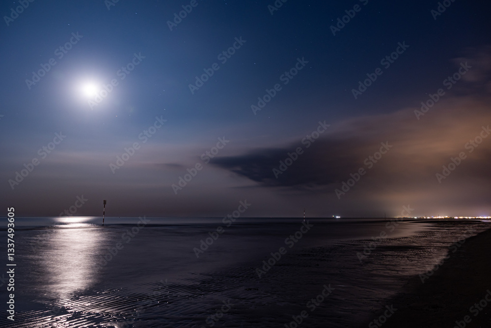 panoramica notturna su un ambiente marino sul mare Adriatico, dalla costa della regione Veneto, con mare calmo, mentre la luna si riflette sull'acqua, in estate, con vista su una città lontana