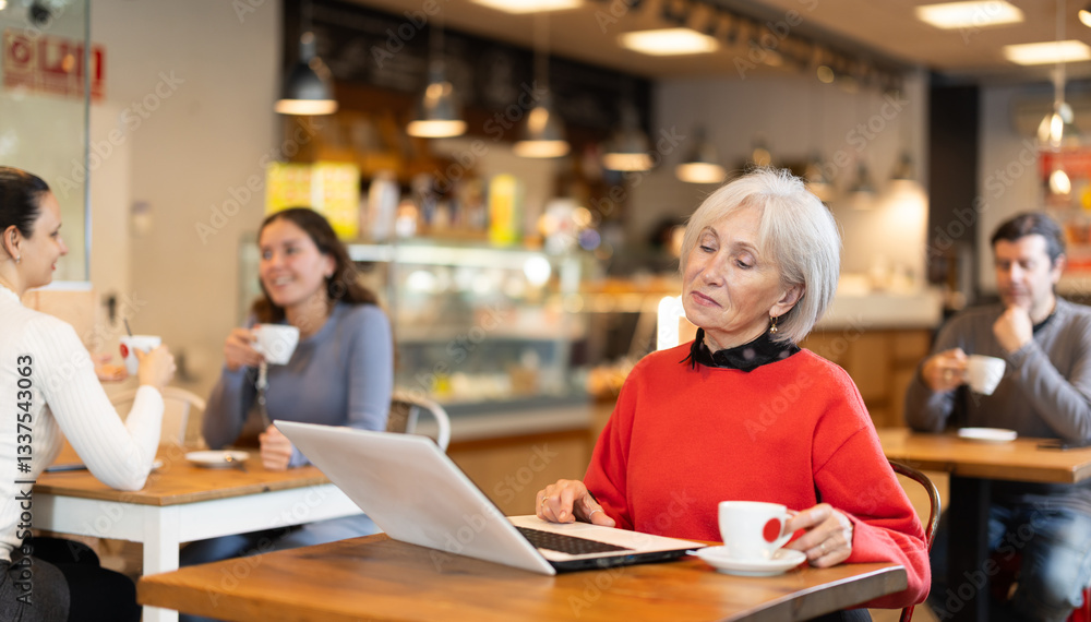 Elderly woman sitting at table, using laptop and enjoing cup of tea in coffee house Elderly woman sitting at table, using laptop and enjoing cup of tea in coffee house