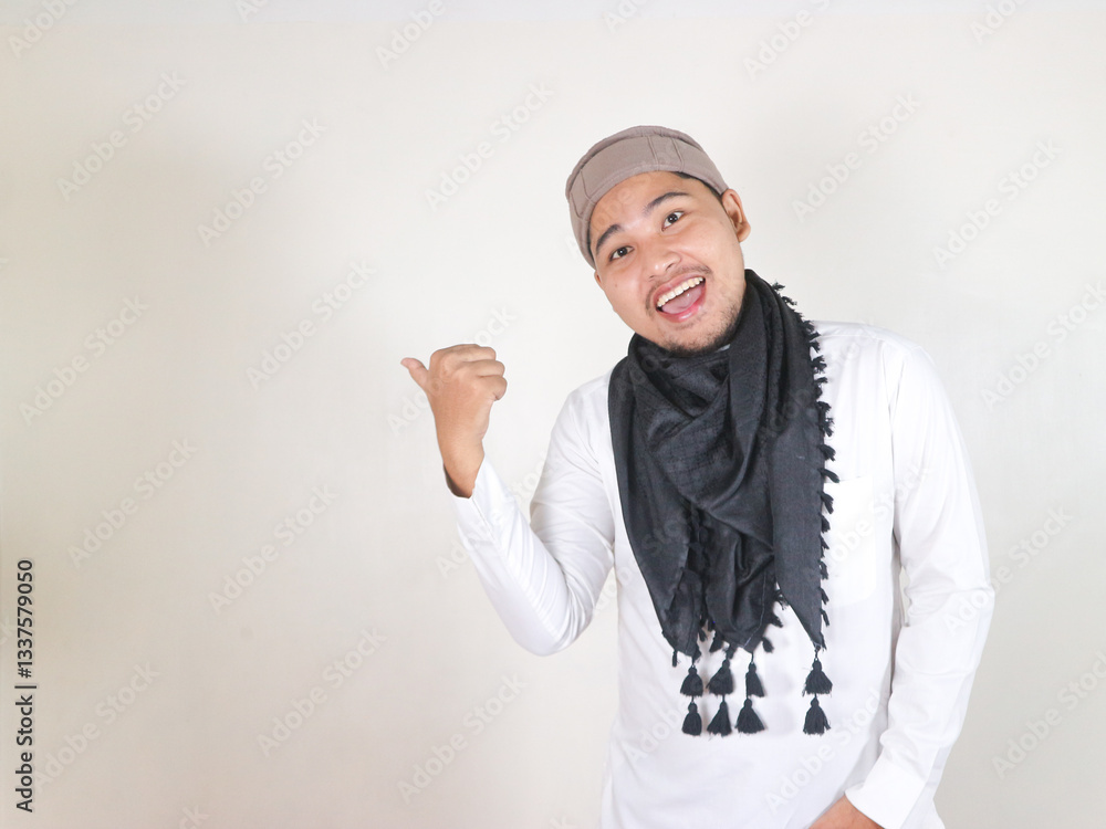 Handsome Asian man in Muslim clothes on white isloted background showing funny expression while pointing to the side Handsome Asian man in Muslim clothes on white isloted background showing funny expression while pointing to the side