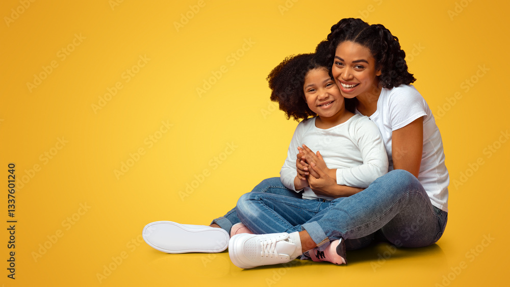 Fototapeta premium Happy Loving Family. Smiling Young African Mother Embracing Her Lovely Little Daughter While Sitting On Floor Over Pink Background Together