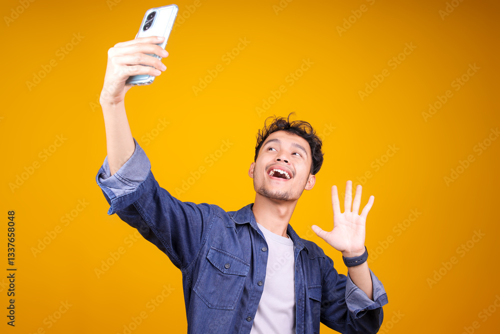 Happy Young Man Video Calling, Talking Online With Mobile Phone, Saying Hello To Smartphone Camera And Waving Hand, Standing Over Yellow Background Happy Young Man Video Calling, Talking Online With Mobile Phone, Saying Hello To Smartphone Camera And Waving Hand, Standing Over Yellow Background