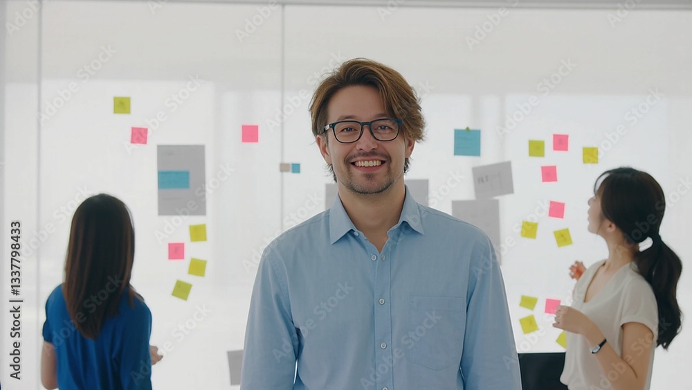 Fototapeta premium Smiling man with glasses stands in a bright office space, colleagues brainstorming with sticky notes.