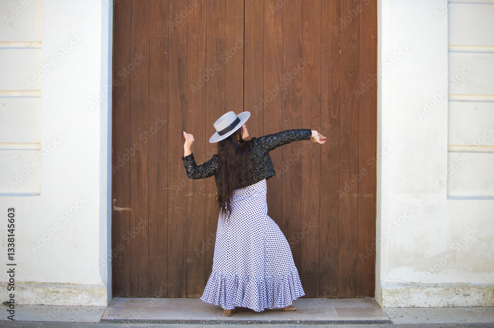 Naklejka premium Latin woman, young and beautiful dancing flamenco in front of a wooden door in a typical street of andalusia. The woman is dressed in a white ruffled dress with black polka dots, a hat and a jacket.