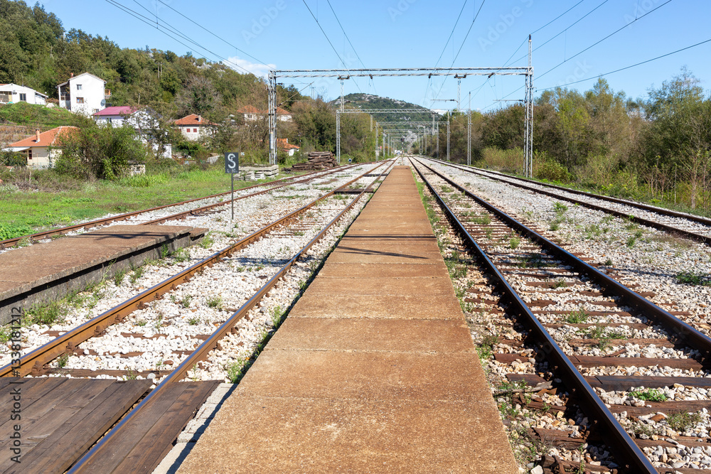 Rural train tracks and empty platform under clear blue sky, surrounded by lush greenery and distant houses Rural train tracks and empty platform under clear blue sky, surrounded by lush greenery and distant houses