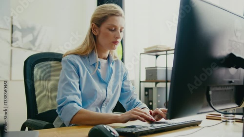 Focused Caucasian woman wearing shirt sitting at desk and typing on keyboard. Concentrating while working on project or responding to corporate emails. Modern workspace with large monitor.