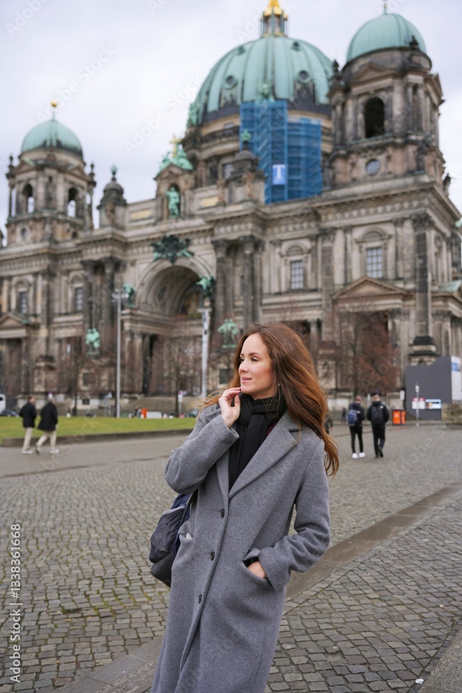 Fototapeta premium A stylish woman in a gray coat and black scarf enjoys a day in Berlin, standing in front of the historic Berlin Cathedral on a cloudy day.
