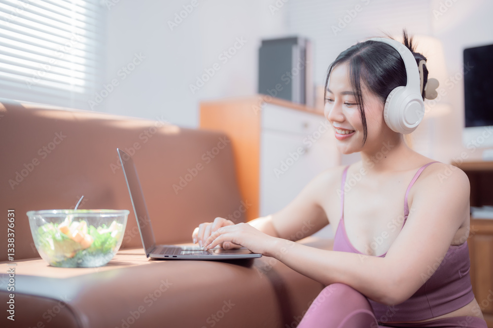 Smiling young sportswoman wearing headphones is using a laptop while sitting on the floor near the sofa, with a bowl of fresh salad next to it, promoting a healthy and connected lifestyle Smiling young sportswoman wearing headphones is using a laptop while sitting on the floor near the sofa, with a bowl of fresh salad next to it, promoting a healthy and connected lifestyle