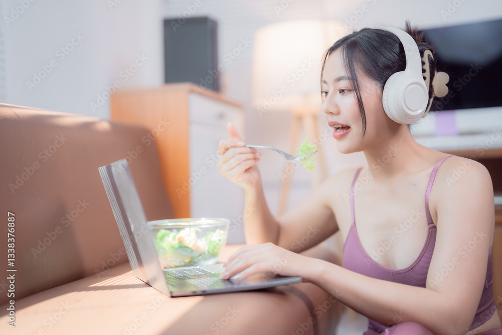 Young sportswoman wearing headphones and sportswear eating a healthy salad while working on her laptop, enjoying a balanced lifestyle in her living room Young sportswoman wearing headphones and sportswear eating a healthy salad while working on her laptop, enjoying a balanced lifestyle in her living room
