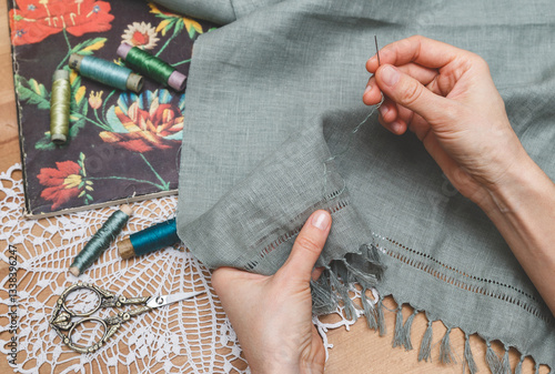 Hands meticulously working on openwork embroidery on grey fabric, surrounded by vintage scissors, thread spools, and a floral book on a lace doily, showcasing a delicate crafting process.