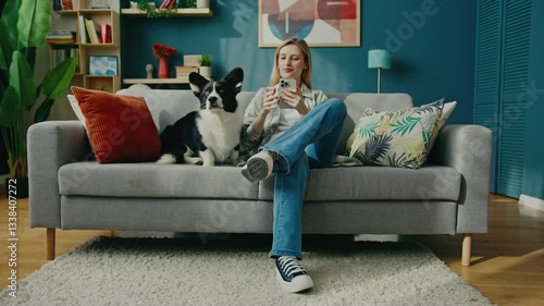 Smiling Caucasian female sitting on modern sofa at home and holding smartphone. Looking at screen while petting beloved dog. Chatting with someone remotely. Corgi resting beside. Enjoying cozy moment.
