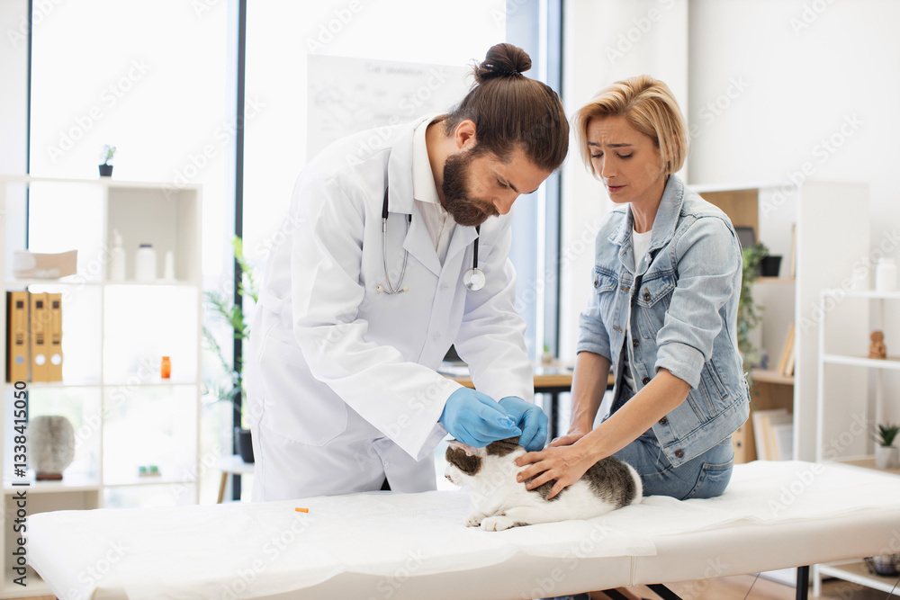 Fototapeta premium Male veterinarian with beard vaccinates cat while blond woman observes in bright clinic setting.