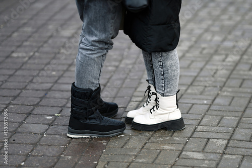 Obraz na płótnie Urban street fashion, close-up of two pairs of stylish boots, one black and one white, symbolizing contrast, elegance, and modern footwear trends. Close-up of two pairs of feet in boots on sidewalk w salonie