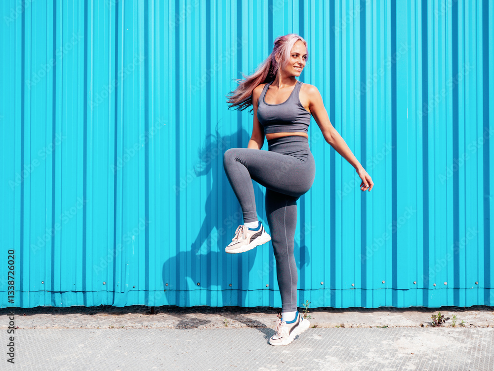 Fototapeta premium Fitness smiling woman in grey sports clothing with pink hair. Young beautiful model with perfect body.Female posing in the street near blue wall.Cheerful and happy. Stretching out before training