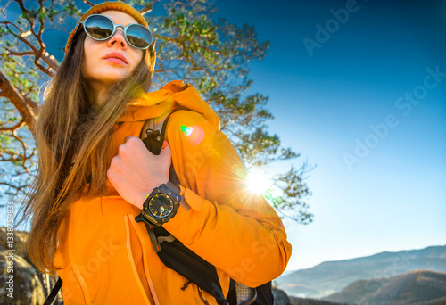 Young woman in orange hiking jacket and sunglasses standing in mountains, holding backpack and wearing wristwatch