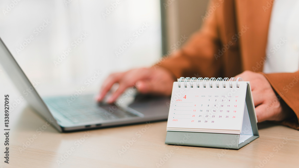 Monthly calendar April 2025 on desk beside professional using laptop, illustrating schedule planning, daily tasks organization, productivity management, and business deadlines.