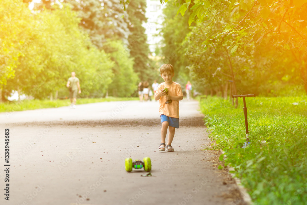 A smiling boy in summer clothes holds a green remote control while squatting on a park path. Outdoor fun with toys on a warm and sunny day. A smiling boy in summer clothes holds a green remote control while squatting on a park path. Outdoor fun with toys on a warm and sunny day.