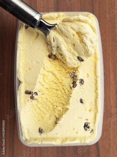 Closeup overhead shot of a freshly opened tub of rum and raisin ice cream, with a full scoop having removed the first spoonful, ready to eat. Use vertically or horizontally.