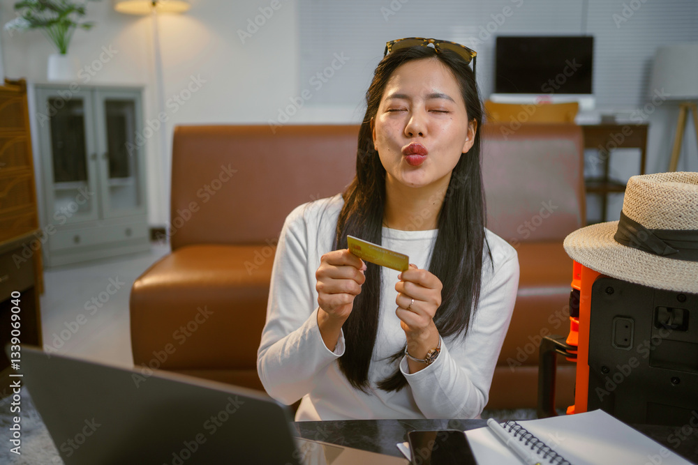 Cheerful traveler kissing credit card near laptop, suitcase beside her, planning upcoming vacation from comfortable home living space Cheerful traveler kissing credit card near laptop, suitcase beside her, planning upcoming vacation from comfortable home living space