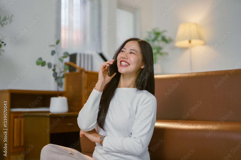 Happy Asian woman chatting on her phone while sitting on the floor near a sofa in a bright, cozy living room Happy Asian woman chatting on her phone while sitting on the floor near a sofa in a bright, cozy living room