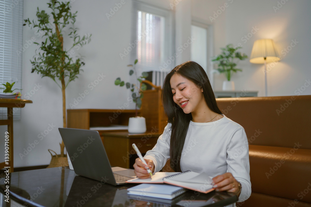 Happy freelancer working remotely using laptop and taking notes in notebook, sitting at desk in cozy living room Happy freelancer working remotely using laptop and taking notes in notebook, sitting at desk in cozy living room