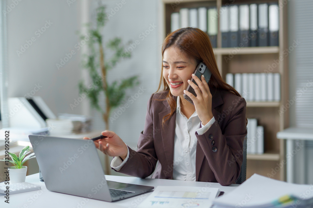 Young Asian manager working on laptop and discussing project with client on mobile phone, sitting at desk in modern office Young Asian manager working on laptop and discussing project with client on mobile phone, sitting at desk in modern office