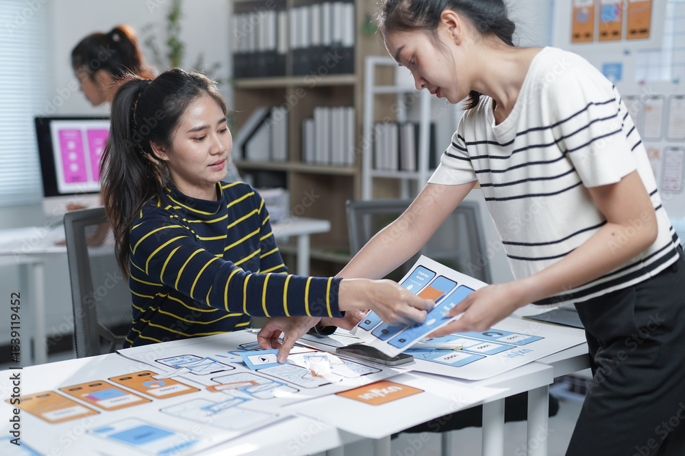 Two Asian female ux designers working together on a mobile app interface wireframe design at a modern bright office Two Asian female ux designers working together on a mobile app interface wireframe design at a modern bright office