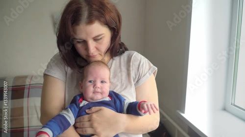 A young mother holds her newborn baby in her arms, stands at the window and cradles him.