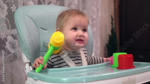 A six-month-old baby sits on a high chair and plays with a toy hammer