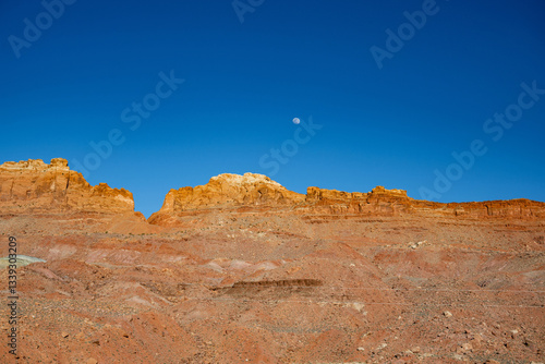 Das Bild zeigt eine weite Wüstenlandschaft unter strahlend blauem Himmel, mit schroffen Bergen und Schluchten im Hintergrund und einem Vollmond, der über dem Horizont aufgeht.