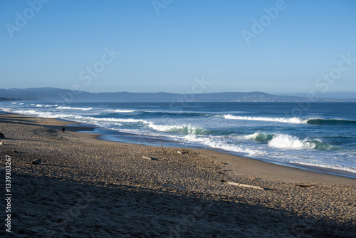 ine ruhige Strandszene mit einem Sandstrand, der unter blauem Himmel sanft ins Meer abfällt. Schaum von Wellen bildet eine Kulisse für den Vordergrund. In der Ferne sind einige Möwen zu sehen